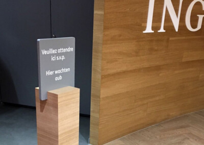 Totem de signalisation intérieure avec le message "Veuillez attendre ici s.v.p Hier wachten aub" dans la banque ING